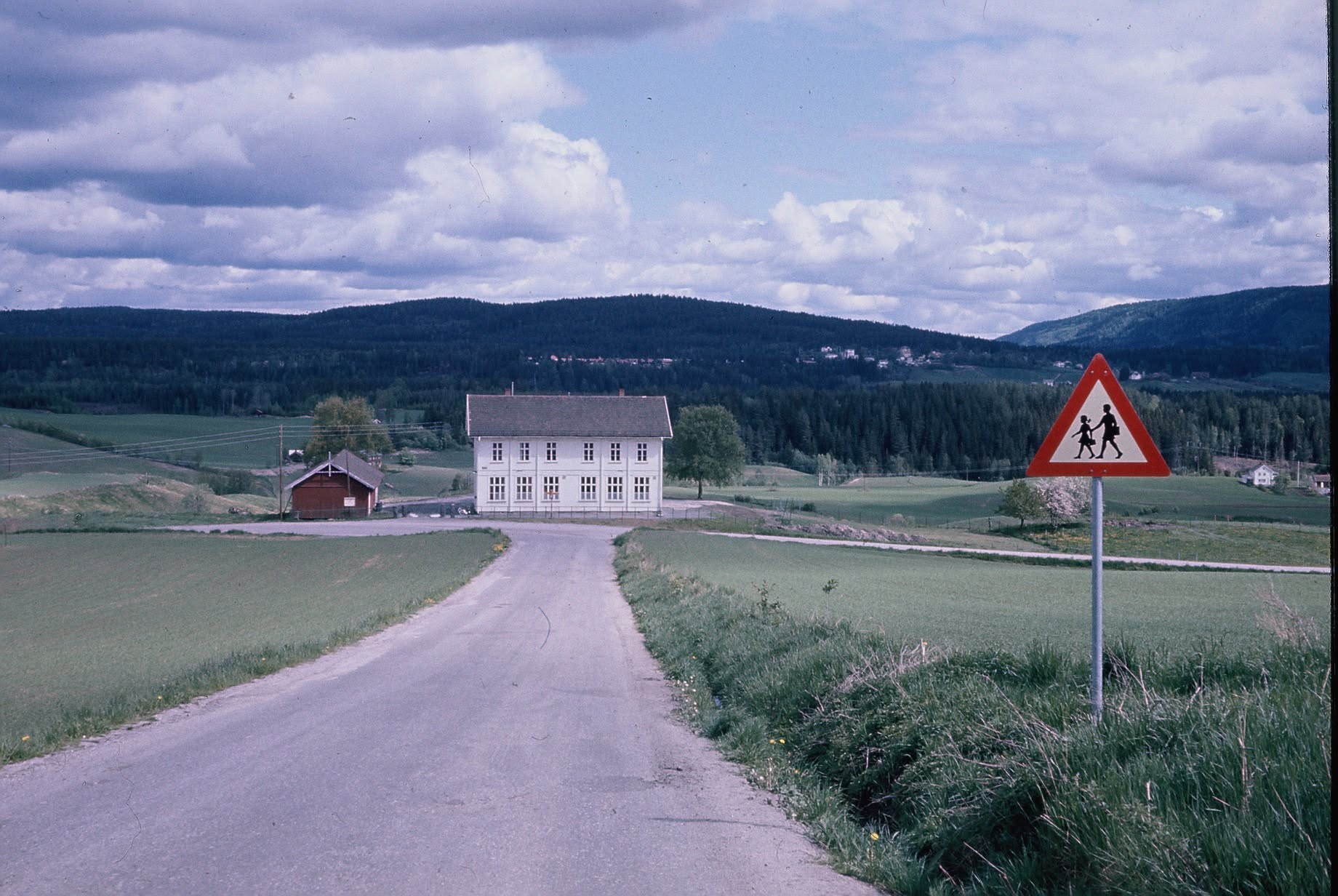Bergbakken (gammel trasé) frem mot det hvite toetasjes skolehuset fra 1907 og det røde sidebygget (stod der SFO bygget står i dag) på Berg Skole i Øvre Sandsvær. Ukjent årstall